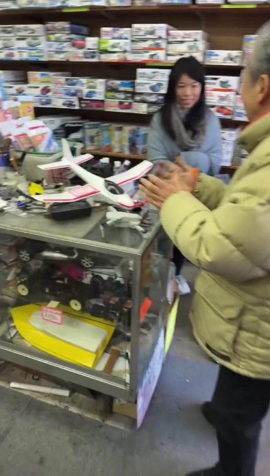 模型店で店員と客が白い飛行機の模型を囲んでいる様子