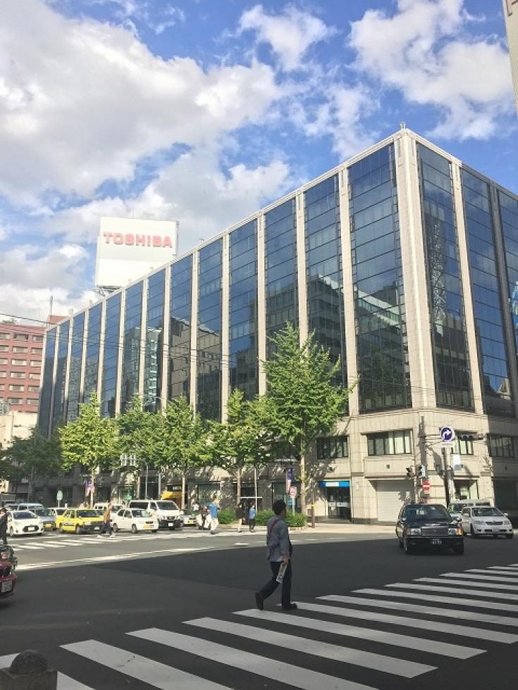 幸田|街角の交差点とガラス張りのビル。青空の下、横断歩道を渡る人々や車。