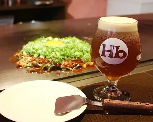 鉄板の上で焼かれる広島焼きと、店のロゴが入ったグラスのビール